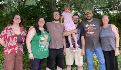 Seven family members of different ages standing together under pine trees, one little girl held up in the middle