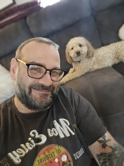 Scott Lutz smiling, with Teddy the cockapoo resting on the couch behind him